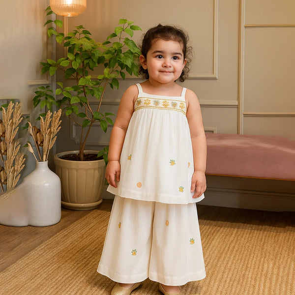 cute girl in a white dress with gold embroidery standing in a room with plants and furniture.