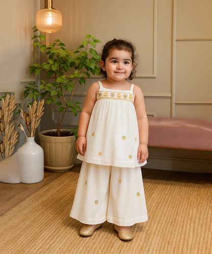 cute girl in a white dress with gold embroidery standing in a room with plants and furniture.