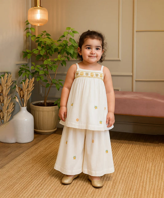 cute girl in a white dress with gold embroidery standing in a room with plants and furniture.