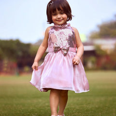 A young girl wearing a pink sleeveless dress with a bow and sequin detail on the top, walking on grass.