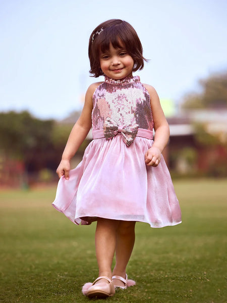 A young girl wearing a pink sleeveless dress with a bow and sequin detail on the top, walking on grass.