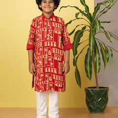 Child wearing a red traditional outfit with white patterns, standing next to a potted plant against a yellow wall.