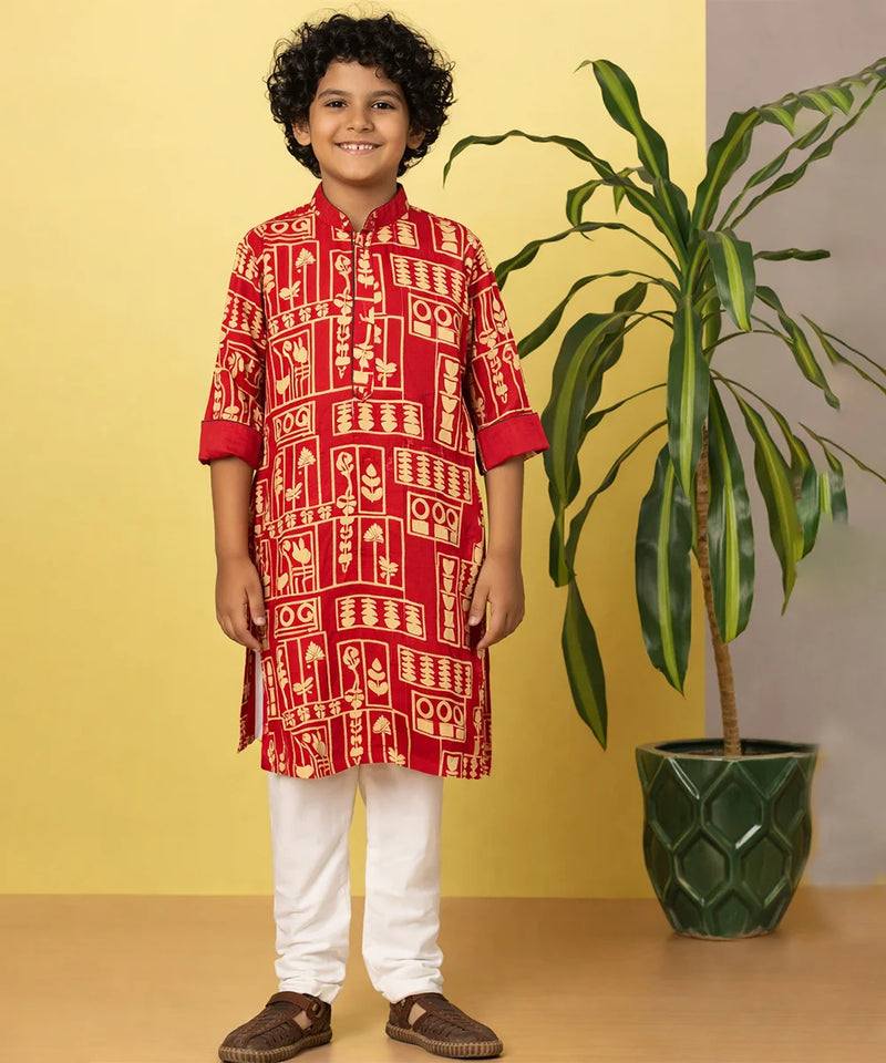 Child wearing a red traditional outfit with white patterns, standing next to a potted plant against a yellow wall.