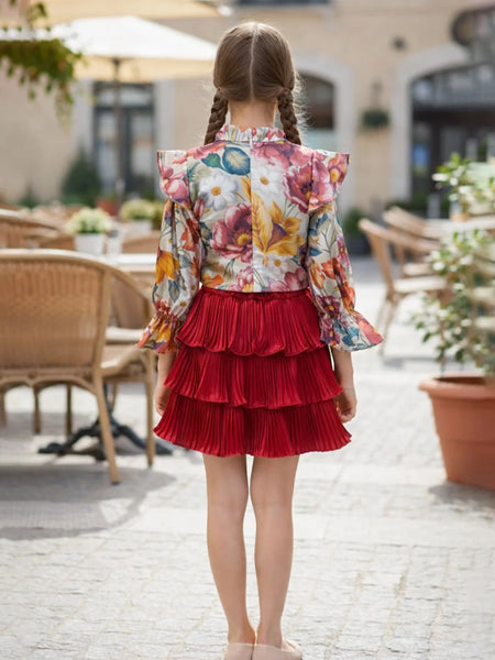 Young girl in a floral top and red skirt standing outdoors on a patio.