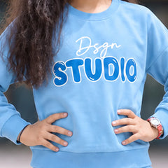 A person wearing a light blue sweatshirt with 'Dsgn STUDIO' printed across the chest in white and blue, paired with matching light blue pants. The person has long dark curly hair and is standing with hands on hips.