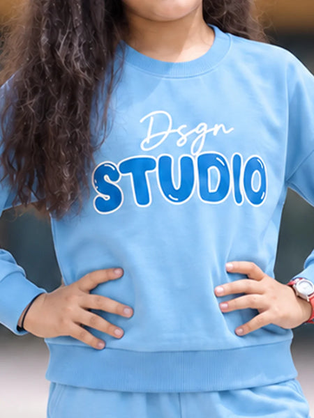A person wearing a light blue sweatshirt with 'Dsgn STUDIO' printed across the chest in white and blue, paired with matching light blue pants. The person has long dark curly hair and is standing with hands on hips.