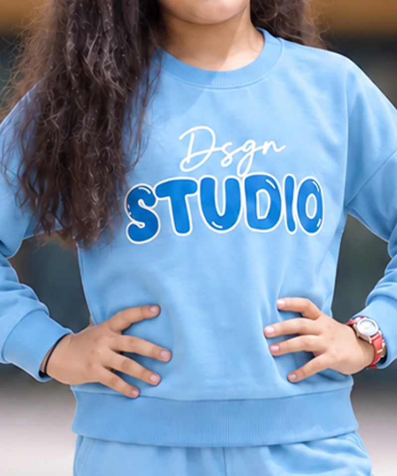 A person wearing a light blue sweatshirt with 'Dsgn STUDIO' printed across the chest in white and blue, paired with matching light blue pants. The person has long dark curly hair and is standing with hands on hips.
