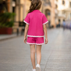 A young girl is shown from behind walking down a blurred city street, wearing a bright pink two-piece pajama or lounge set with white scalloped trim on the short-sleeve top and shorts.