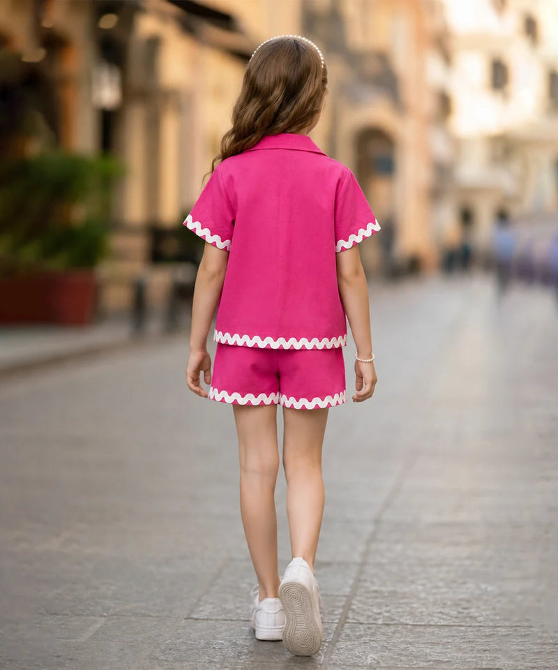 A young girl is shown from behind walking down a blurred city street, wearing a bright pink two-piece pajama or lounge set with white scalloped trim on the short-sleeve top and shorts.