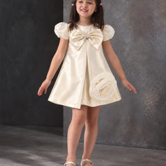 A young girl with dark hair in pigtails models a cream satin flower girl dress with puff sleeves, a large bow at the chest, and a fabric rose detail on the skirt, paired with white shoes against a textured gray studio backdrop.