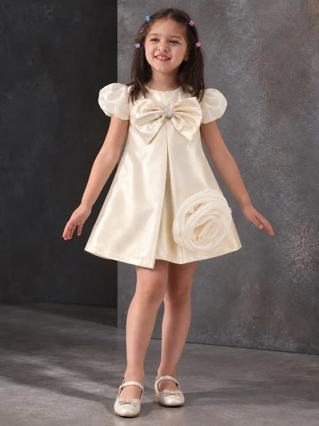 A young girl with dark hair in pigtails models a cream satin flower girl dress with puff sleeves, a large bow at the chest, and a fabric rose detail on the skirt, paired with white shoes against a textured gray studio backdrop.