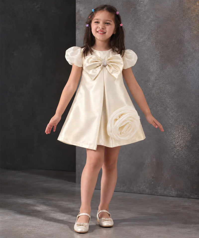 A young girl with dark hair in pigtails models a cream satin flower girl dress with puff sleeves, a large bow at the chest, and a fabric rose detail on the skirt, paired with white shoes against a textured gray studio backdrop.