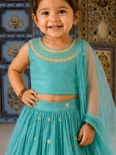 A young child wearing a turquoise blue traditional outfit with gold embroidery, featuring a sleeveless crop top, matching skirt, and sheer dupatta, posed in front of a decorative patterned wall.