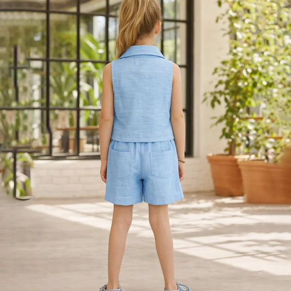 Person wearing a light blue outfit standing in an outdoor setting with plants and a building in the background.