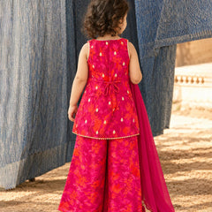 A young child viewed from behind wearing a bright pink and orange floral printed traditional outfit with a matching dupatta, standing outdoors between blue hanging textiles.