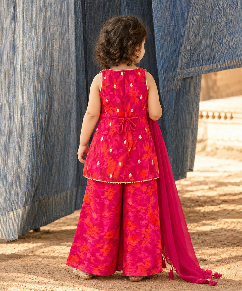 A young child viewed from behind wearing a bright pink and orange floral printed traditional outfit with a matching dupatta, standing outdoors between blue hanging textiles.