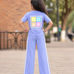 A young girl viewed from behind wearing a light blue two-piece tracksuit with a colorful tic-tac-toe graphic on the back and the words 'Play Station' written below it. She is standing on a paved outdoor walkway with a brick wall and trees in the background.
