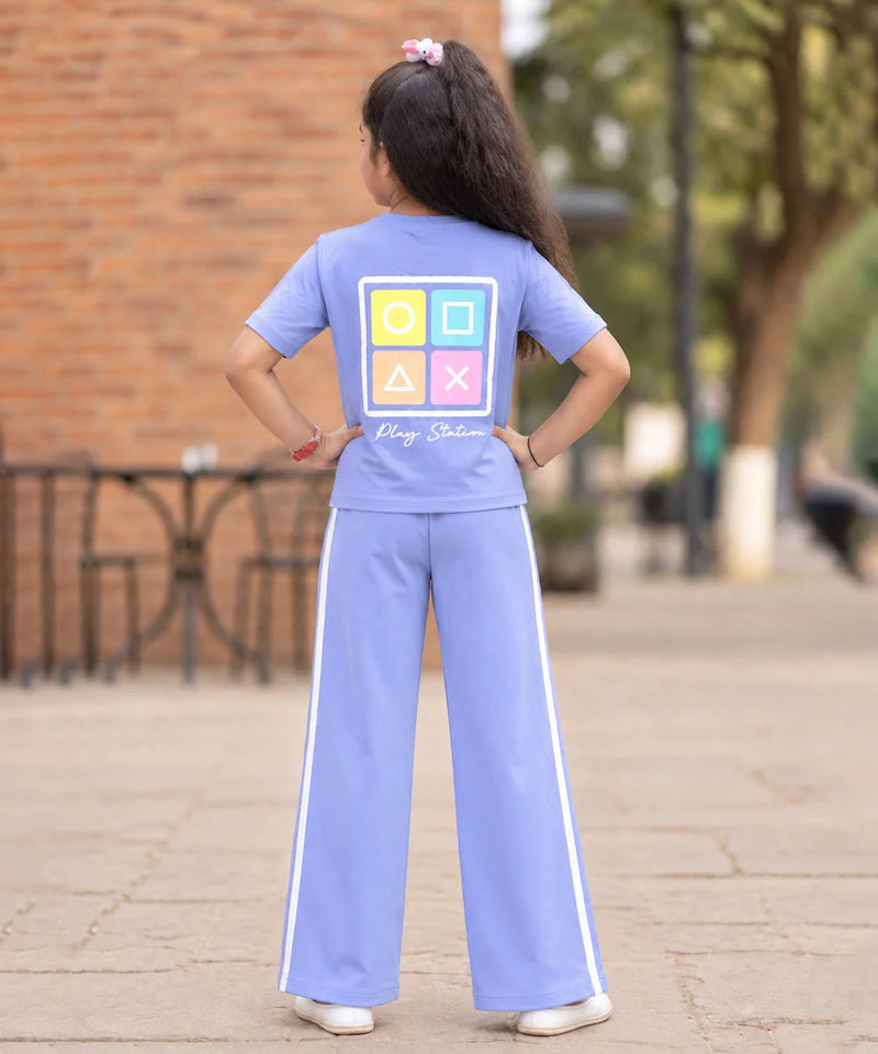 A young girl viewed from behind wearing a light blue two-piece tracksuit with a colorful tic-tac-toe graphic on the back and the words 'Play Station' written below it. She is standing on a paved outdoor walkway with a brick wall and trees in the background.