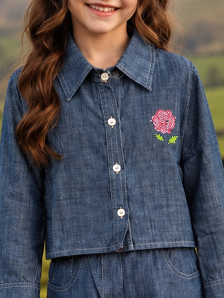 A child wearing a cropped blue denim button-up shirt with a pink rose and green leaf embroidery on the chest, set against a blurred outdoor background.