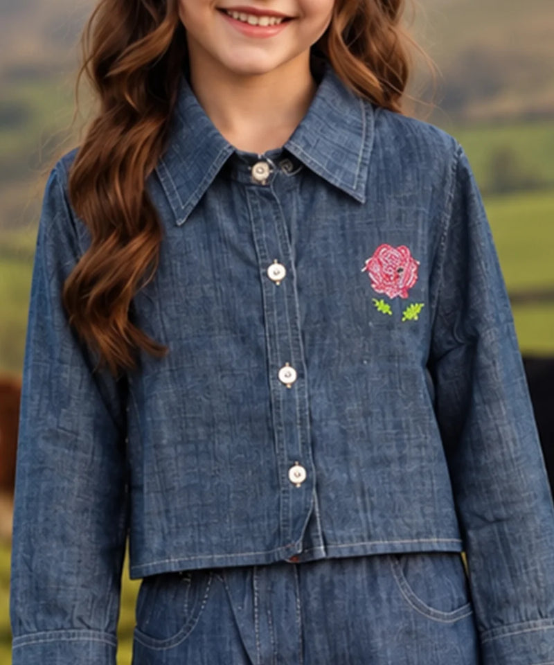 A child wearing a cropped blue denim button-up shirt with a pink rose and green leaf embroidery on the chest, set against a blurred outdoor background.