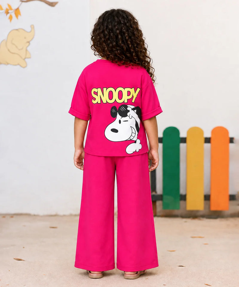 A child wearing a bright pink short-sleeve T-shirt with a large Snoopy graphic on the front. The graphic shows Snoopy wearing black sunglasses, a blue jacket, a yellow shirt, and blue jeans, with the word 'SNOOPY' printed below in yellow.