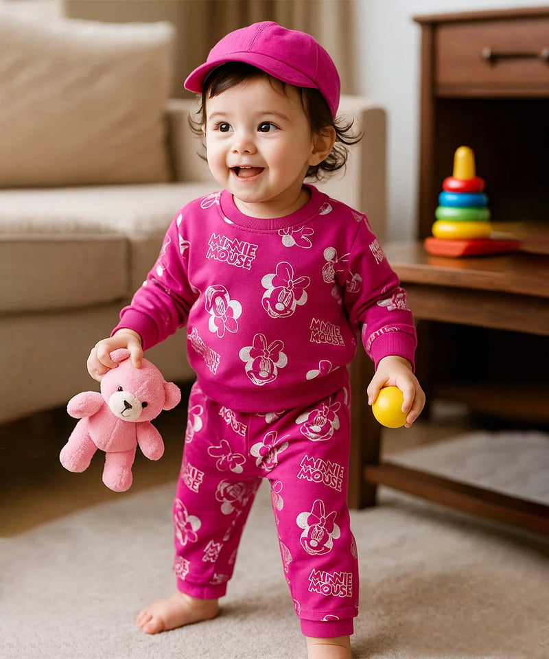 Child wearing a pink Minnie Mouse outfit holding a teddy bear and a ball in a living room.