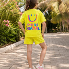 A child wearing a bright yellow matching t-shirt and shorts set, viewed from behind. The shirt features a large colorful smiley face and the text 'SOMETHING DEAR ME' in rainbow colors.