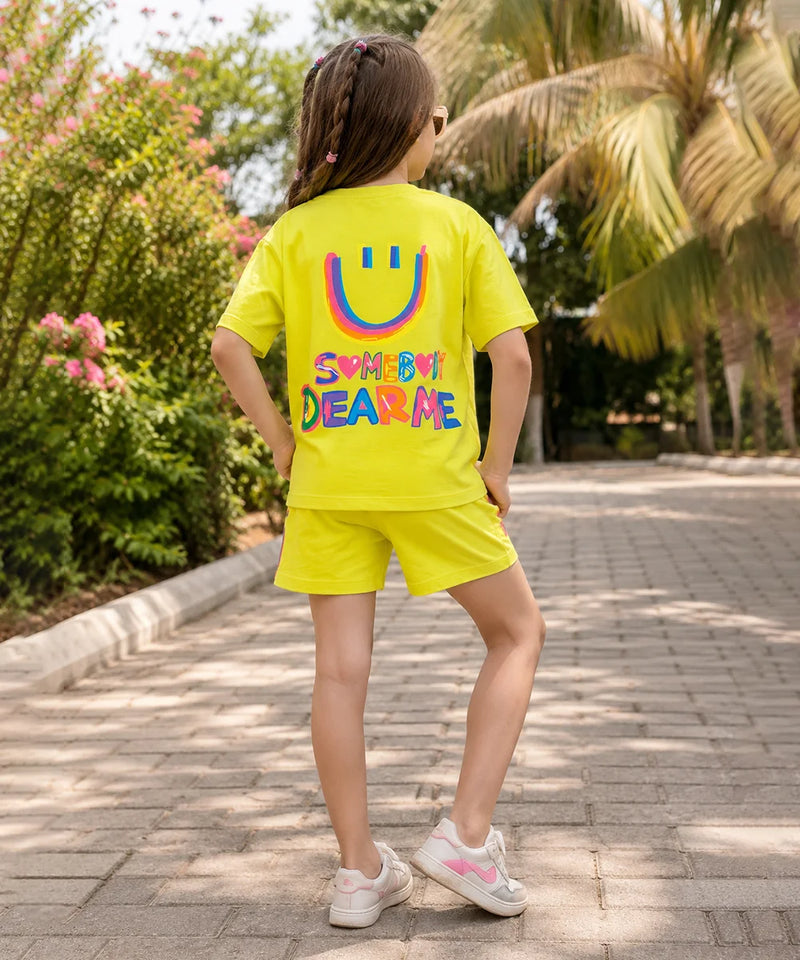 A child wearing a bright yellow matching t-shirt and shorts set, viewed from behind. The shirt features a large colorful smiley face and the text 'SOMETHING DEAR ME' in rainbow colors.