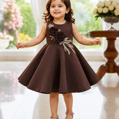 A young girl with curly brown hair stands smiling in a sleeveless dark brown dress with a flared skirt. The dress features a decorative bodice with large dark brown fabric roses and metallic gold leaf appliques.
