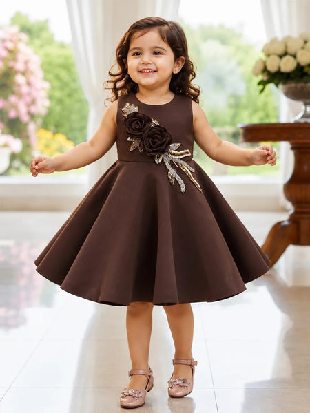 A young girl with curly brown hair stands smiling in a sleeveless dark brown dress with a flared skirt. The dress features a decorative bodice with large dark brown fabric roses and metallic gold leaf appliques.