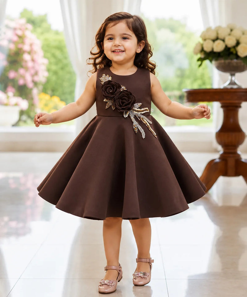 A young girl with curly brown hair stands smiling in a sleeveless dark brown dress with a flared skirt. The dress features a decorative bodice with large dark brown fabric roses and metallic gold leaf appliques.