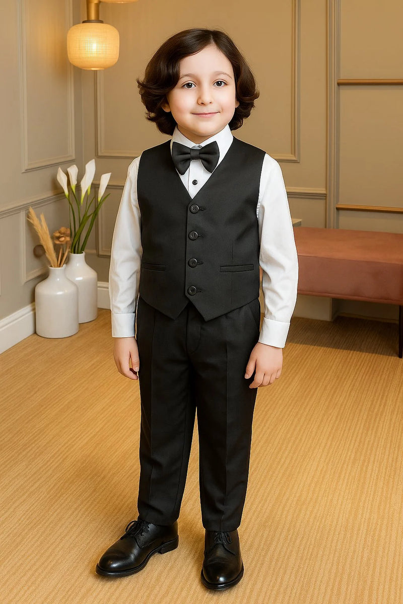Young boy in a formal black suit standing in a room with light wood flooring and neutral walls.