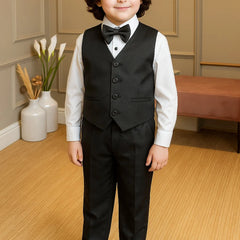 Young boy in a formal black suit standing in a room with light wood flooring and neutral walls.