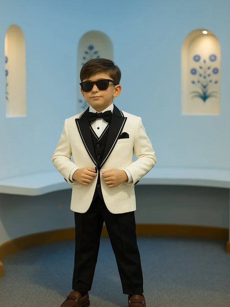 Young boy in a formal tuxedo with sunglasses standing against a light blue wall.