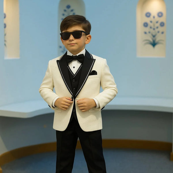 Young boy in a formal tuxedo with sunglasses standing against a light blue wall.