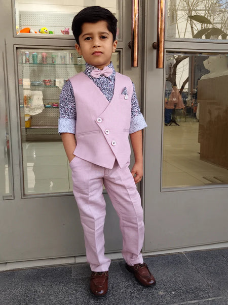 A boy wearing a pink waist coat set with a patterned shirt and trousers, standing in front of a glass door.