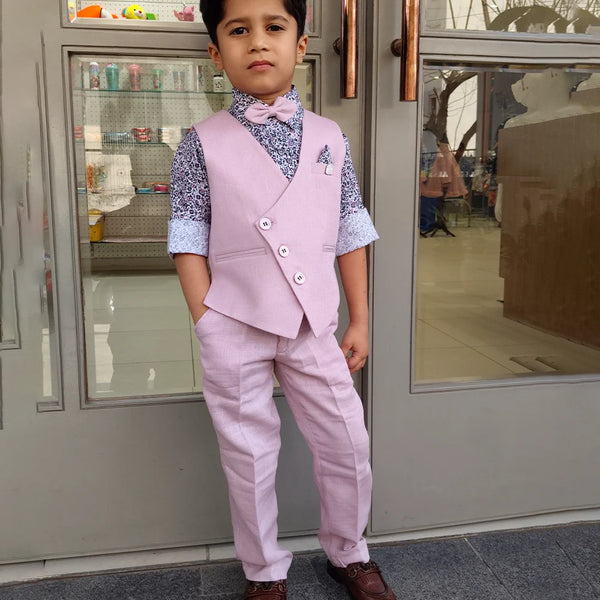 A boy wearing a pink waist coat set with a patterned shirt and trousers, standing in front of a glass door.