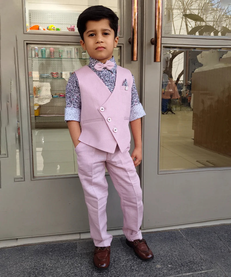 A boy wearing a pink waist coat set with a patterned shirt and trousers, standing in front of a glass door.