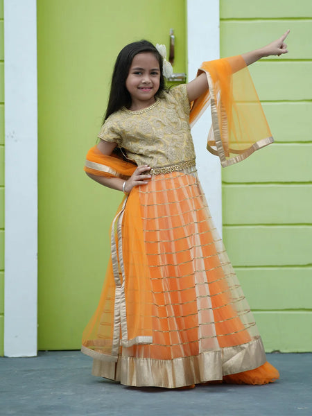 Young girl in an orange and gold traditional outfit standing against a green wall.