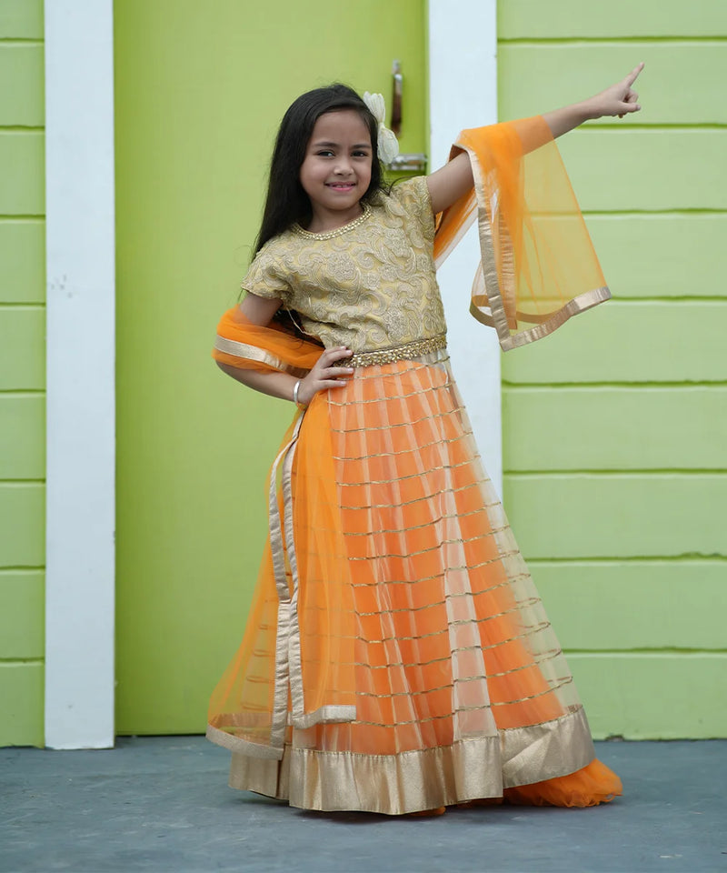 Young girl in an orange and gold traditional outfit standing against a green wall.