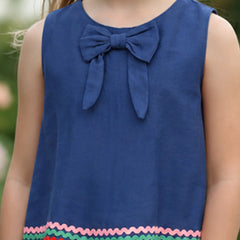 A sleeveless blue children's top with a large bow at the neckline and a colorful zigzag trim along the hem, shown on a child outdoors.