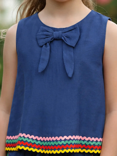 A sleeveless blue children's top with a large bow at the neckline and a colorful zigzag trim along the hem, shown on a child outdoors.