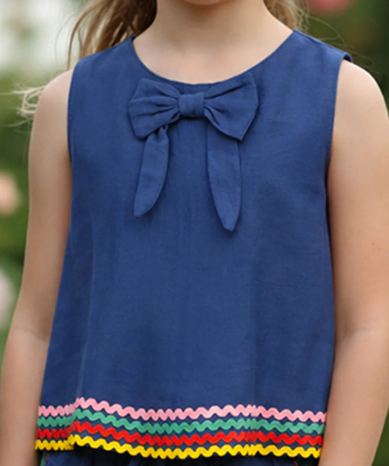 A sleeveless blue children's top with a large bow at the neckline and a colorful zigzag trim along the hem, shown on a child outdoors.