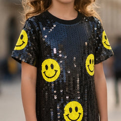 A child wearing a short-sleeve black sequined top covered with yellow smiley face patches, shown from the shoulders to the hips against a blurred outdoor background.