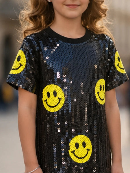 A child wearing a short-sleeve black sequined top covered with yellow smiley face patches, shown from the shoulders to the hips against a blurred outdoor background.