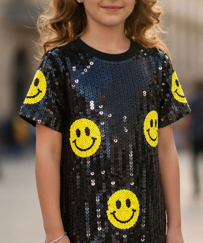 A child wearing a short-sleeve black sequined top covered with yellow smiley face patches, shown from the shoulders to the hips against a blurred outdoor background.