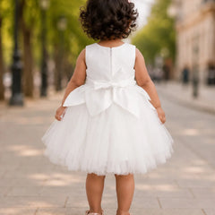 A toddler girl viewed from behind wearing a sleeveless white tulle dress with a large bow at the back, standing on a paved street with blurred trees and buildings in the background.