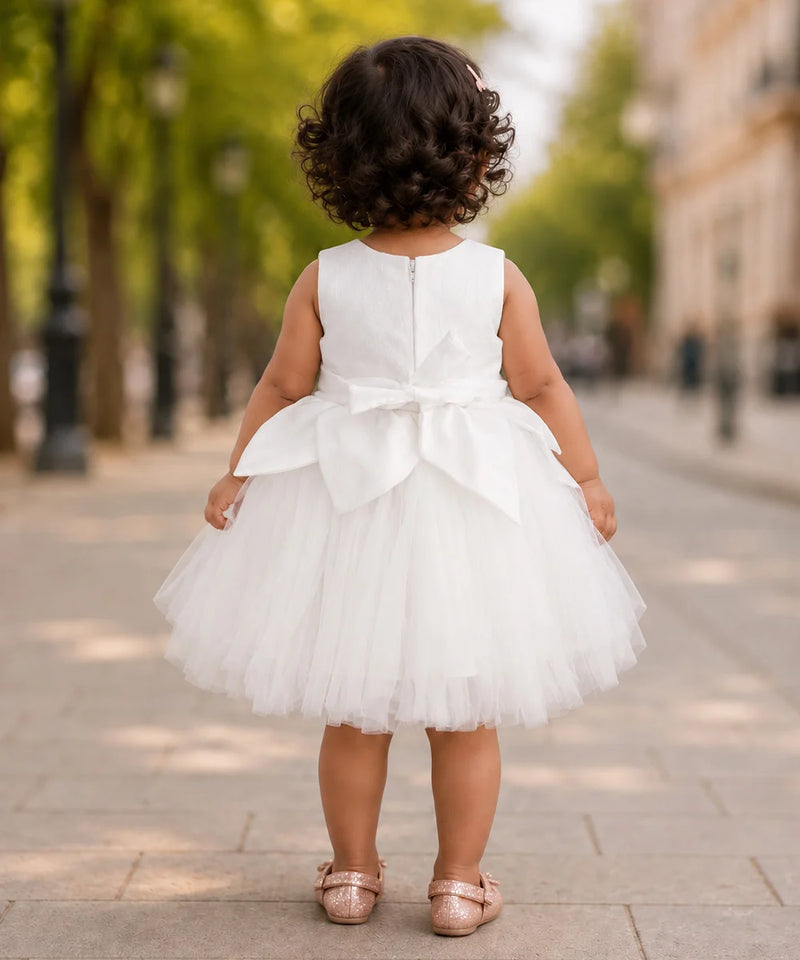 A toddler girl viewed from behind wearing a sleeveless white tulle dress with a large bow at the back, standing on a paved street with blurred trees and buildings in the background.