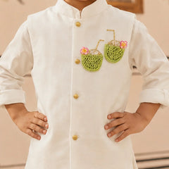 A child wearing a white traditional-style jacket with gold buttons and embroidered green coconut motifs with pink flowers on the chest, standing with hands on hips.