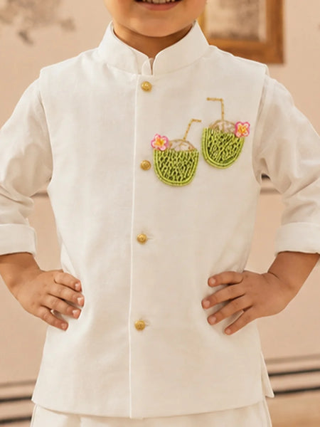 A child wearing a white traditional-style jacket with gold buttons and embroidered green coconut motifs with pink flowers on the chest, standing with hands on hips.